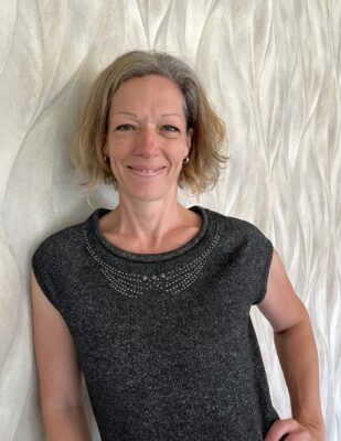 Weisse Frau im mittleren Alters mit blonden, halblangen Haaren und einem schwarzen T-Shirt. Sie ist Mitglied in einem Business-Netzwerk. Sie lehnt mit dem Oberkörper an einer Wand 
