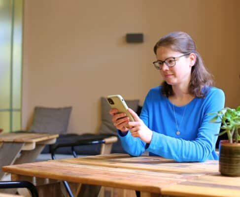 Eine junge Frau mit halblangen Haaren, Brille und blauem Shirt sitzt an einem Tisch mit einem Handy in der Hand.
