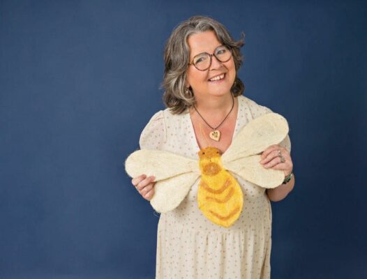 Frau im mittleren Alter mit Brille und in einem hellen Sommerkleid. Sie hält eine Hummel aus Papier in den Händen. 