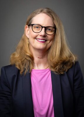 Portrait einer blonden Frau im mittleren Alter mit Brille, rosafarbenem Shirt und blauem Blazer. Sie ist Mentorin für achtsame Selbstführung